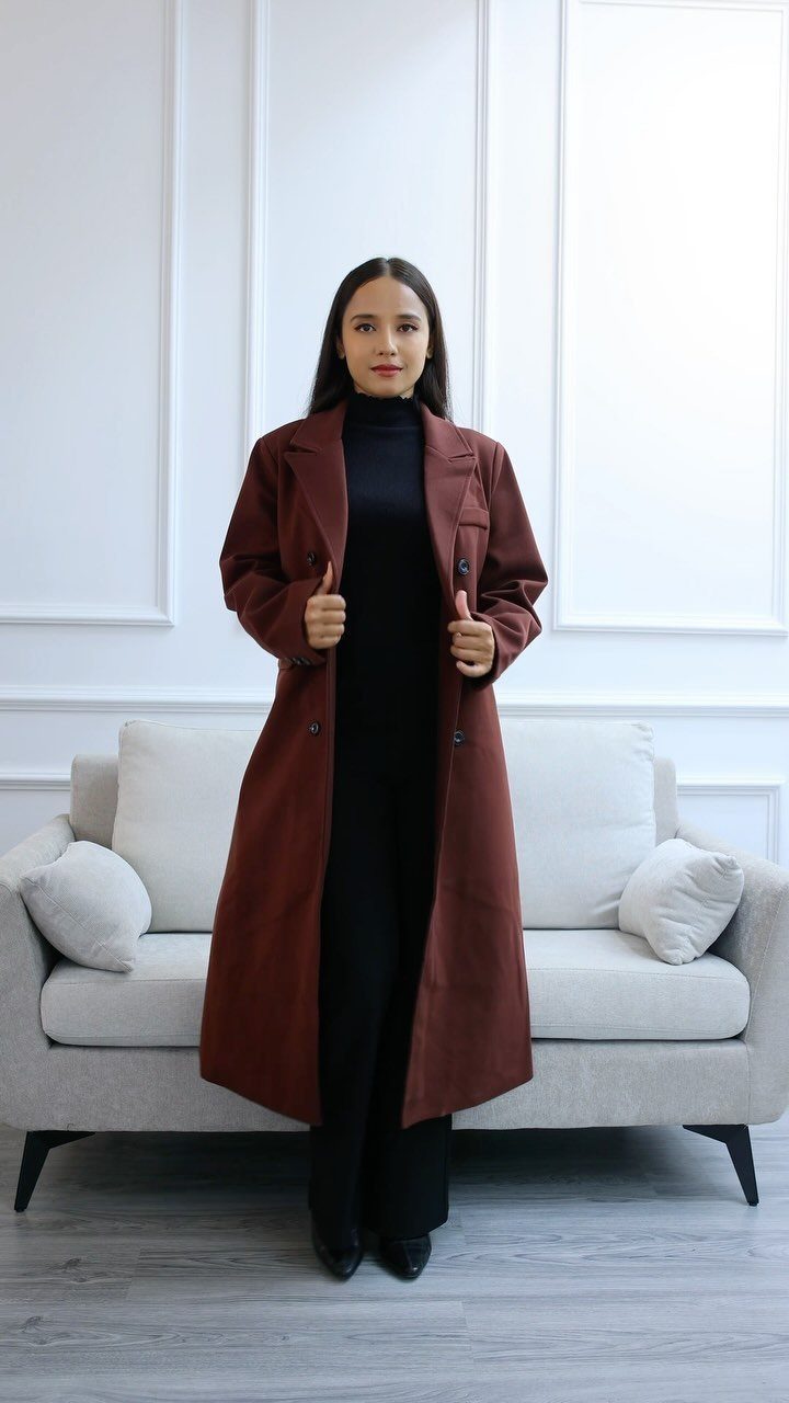 A woman wearing a long reddish-brown wool coat and black turtleneck standing in a bright room.