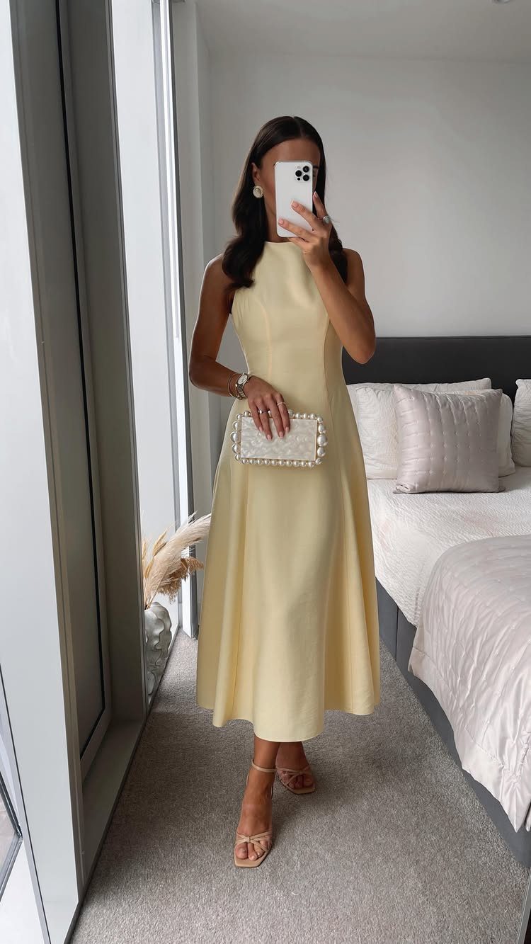 Woman in a pale yellow sleeveless midi dress posing for a mirror selfie in a bedroom.