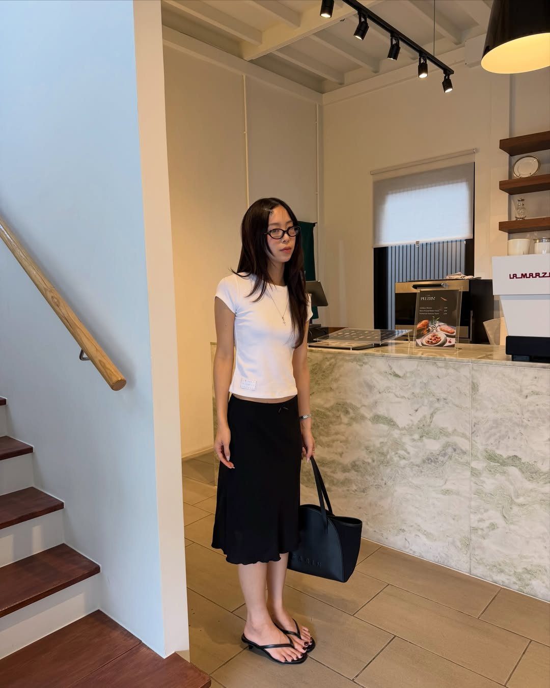 A woman wearing a white top and black midi skirt standing in a bright room.