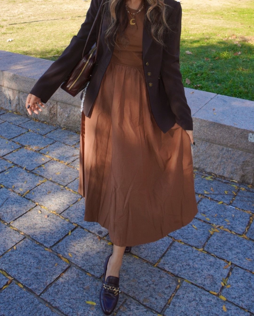 A person wearing a brown midi dress, dark blazer, and loafers standing on a stone path.