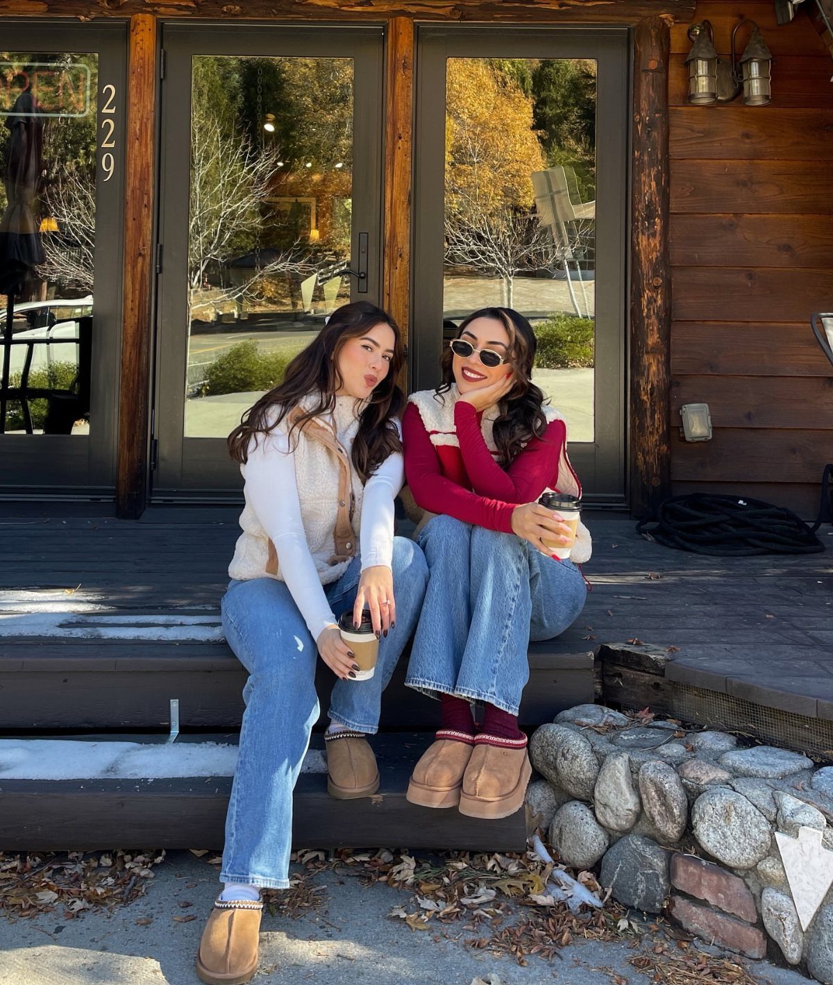 Two women sitting on wooden steps wearing cozy autumn outfits and holding coffee cups.