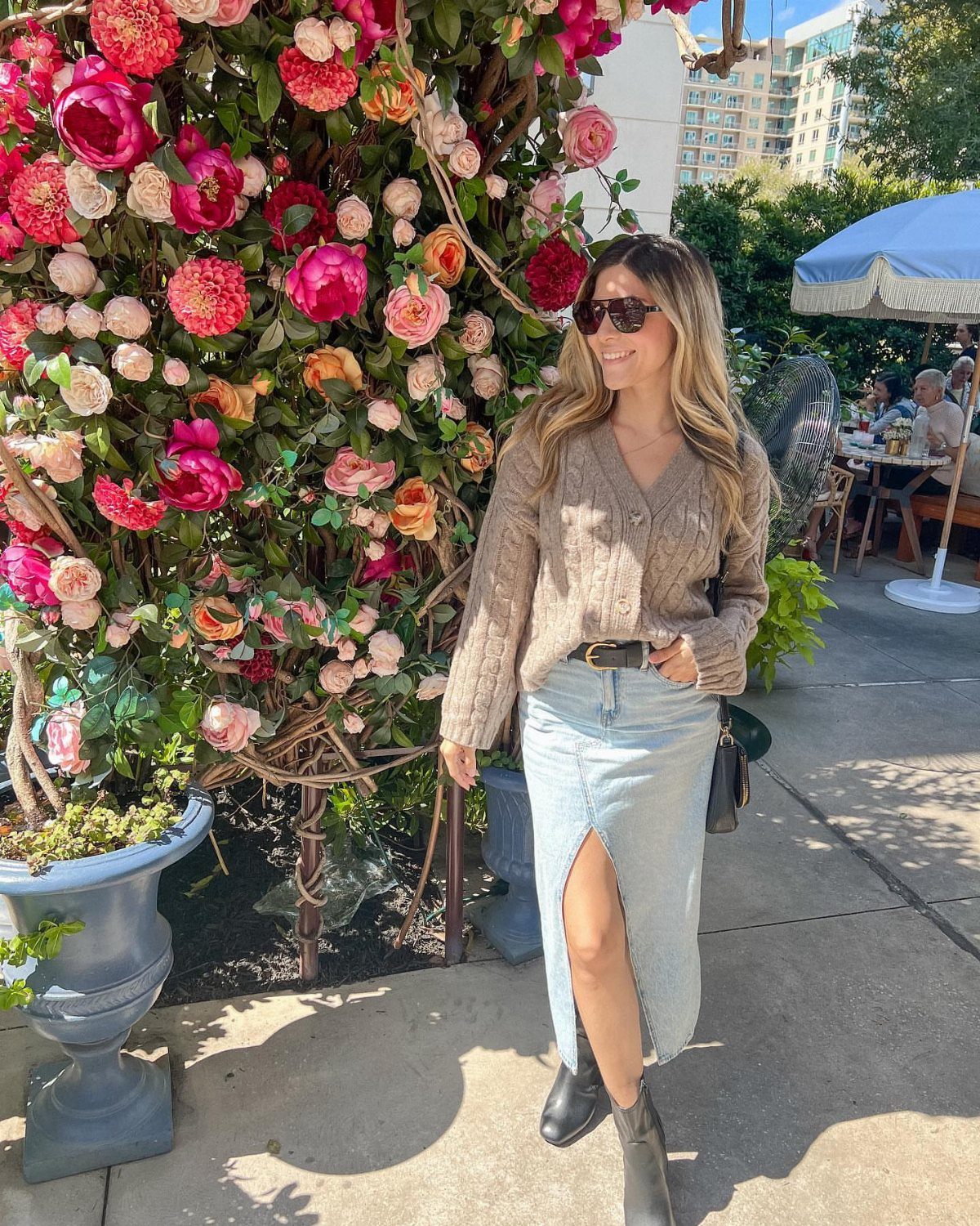 A woman in a beige knit sweater and denim slit skirt standing by pink roses.