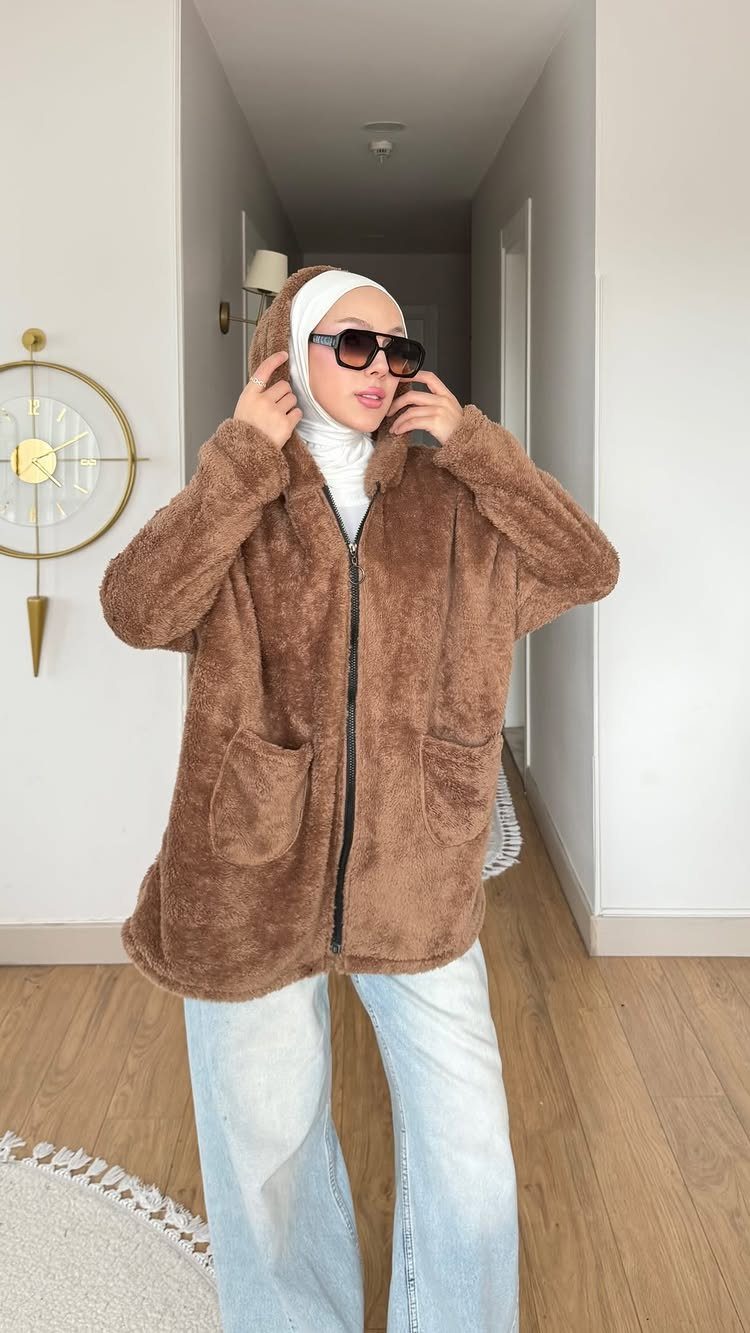 A woman in a brown fuzzy hooded jacket, sunglasses, and light blue jeans standing in a hallway.