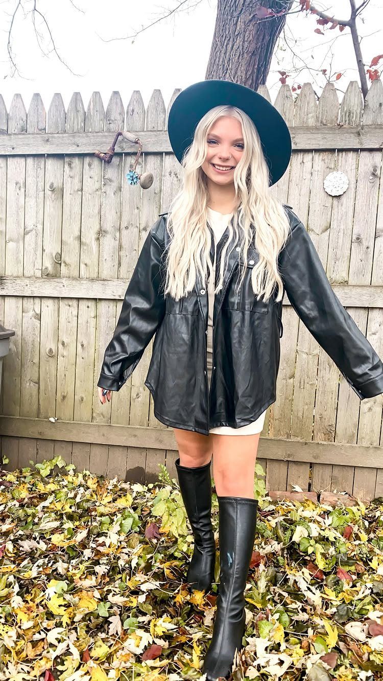 A smiling blonde woman in a black leather jacket and boots standing among autumn leaves.