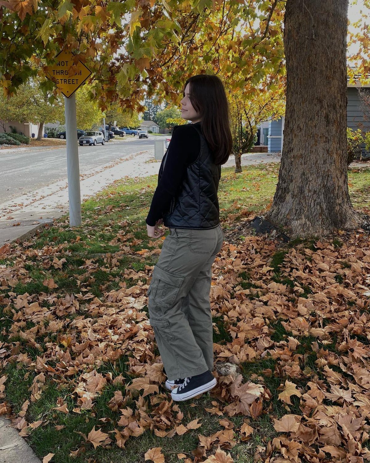 A woman in a black vest and olive cargo pants stands among autumn leaves.