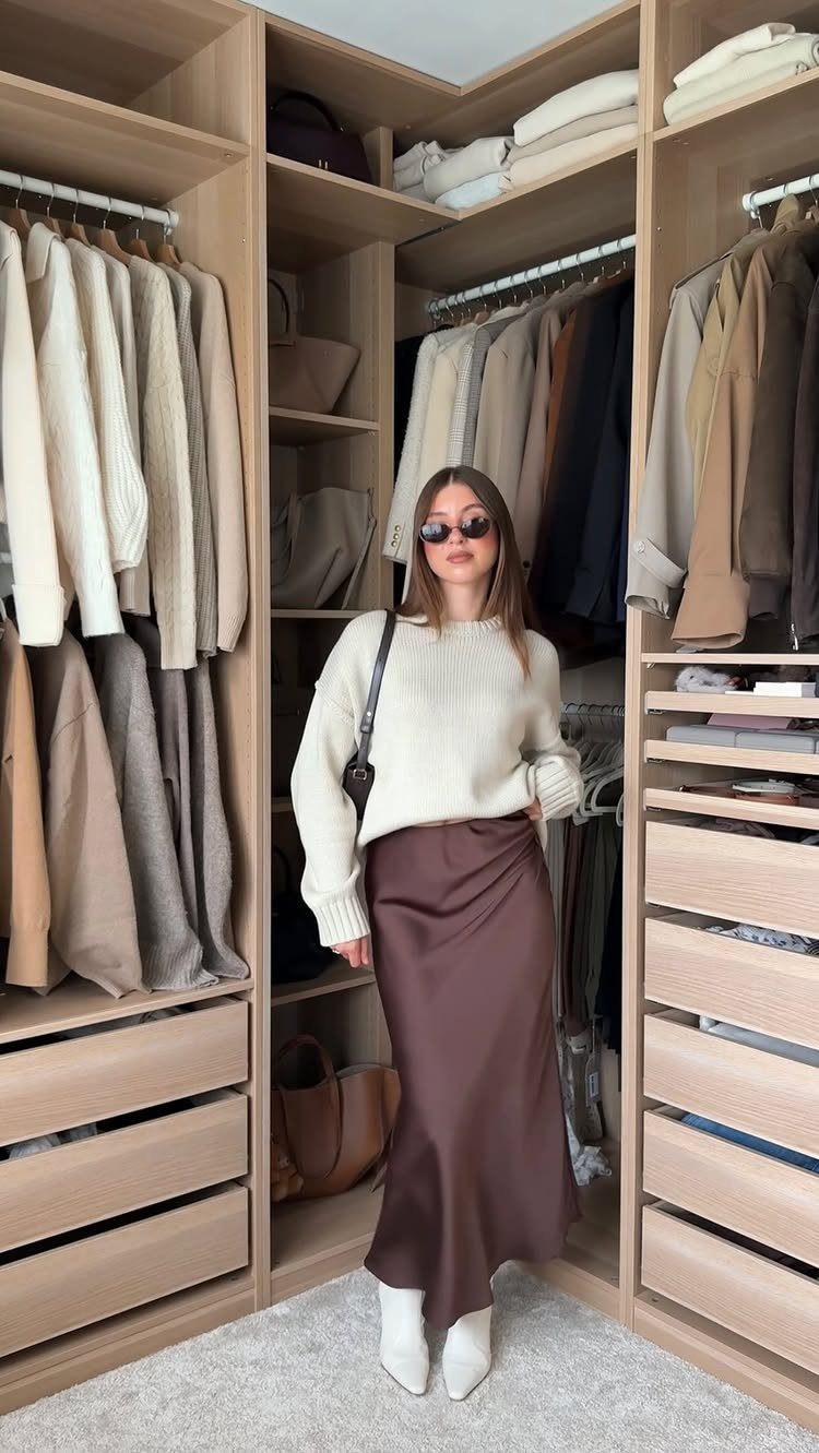 Woman posing in a cream sweater and brown satin maxi skirt inside a large closet.