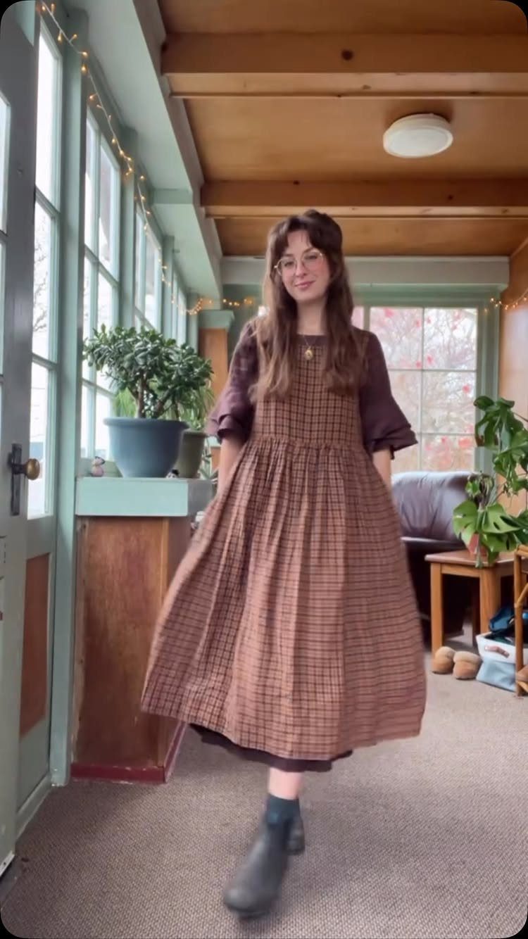 A smiling woman wearing a brown plaid midi dress and black boots standing in a sunlit room.