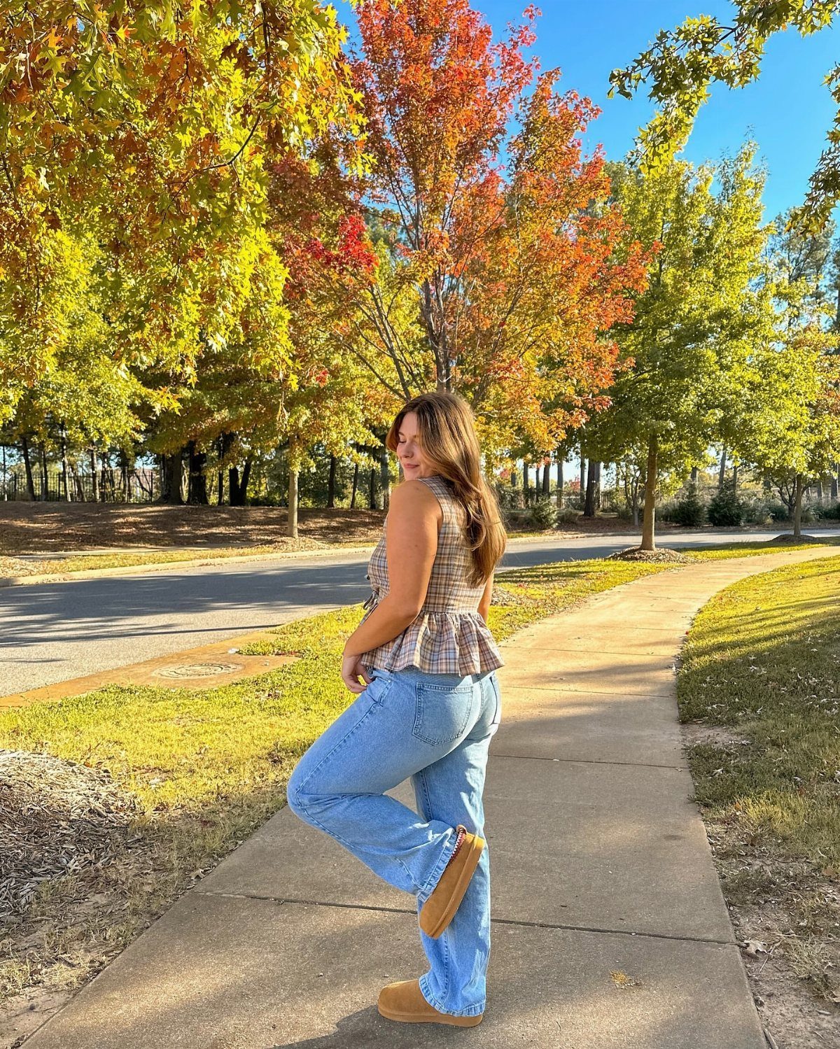 A woman in a plaid peplum top and blue jeans posing on a sidewalk near autumn trees.