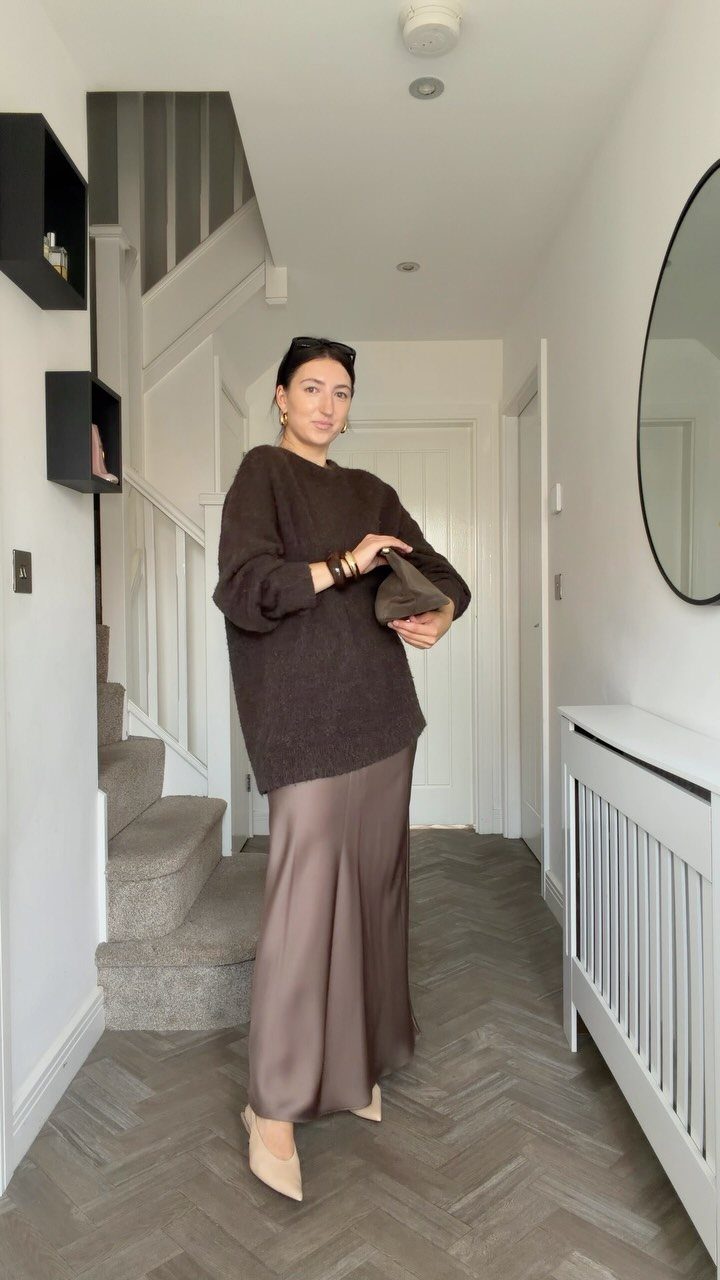 Woman wearing a brown chunky knit sweater and a taupe satin midi skirt in a hallway.