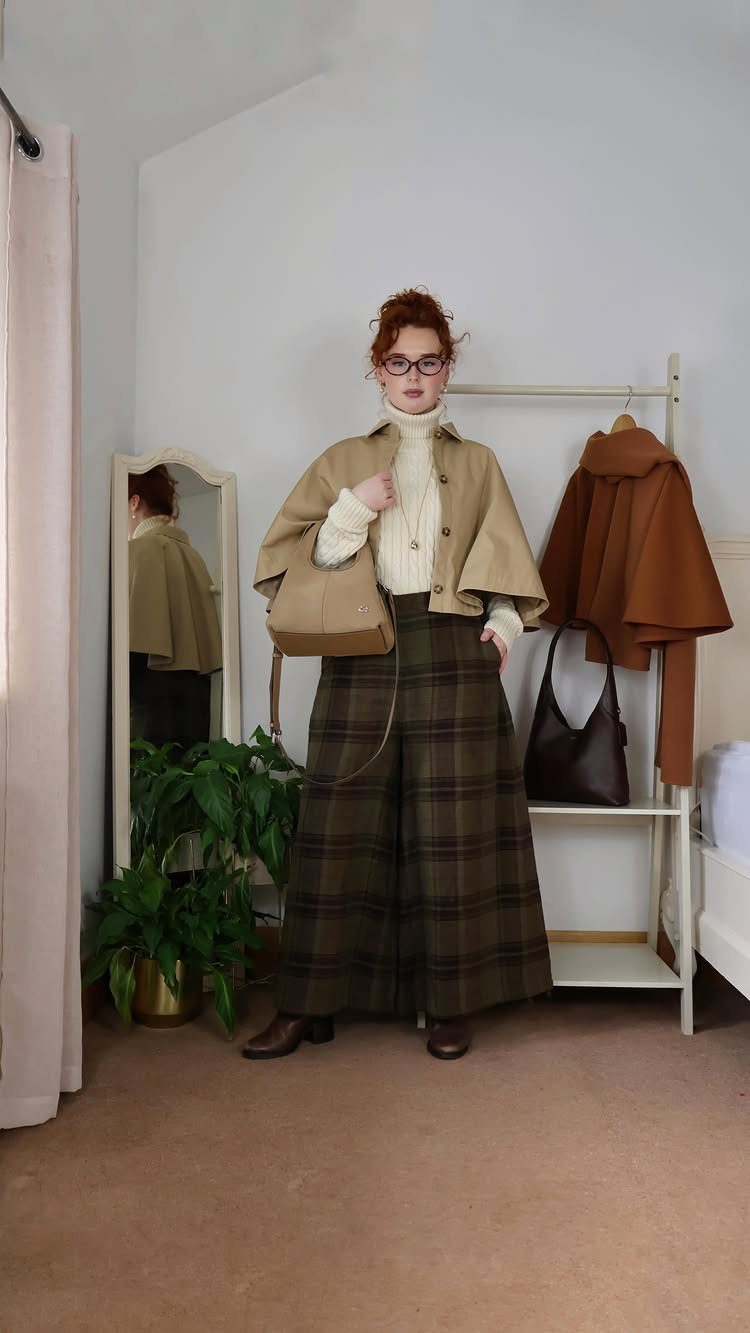 Woman wearing a tan capelet and wide-leg plaid trousers standing in a room.