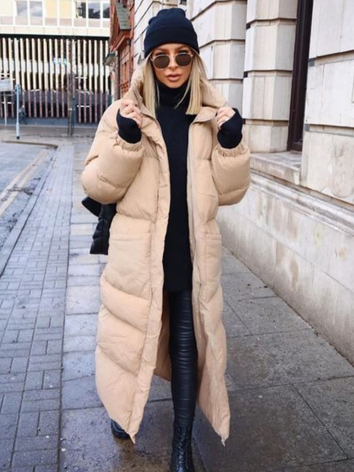 Woman in a long beige puffer coat, black turtleneck, and leather pants standing outdoors.