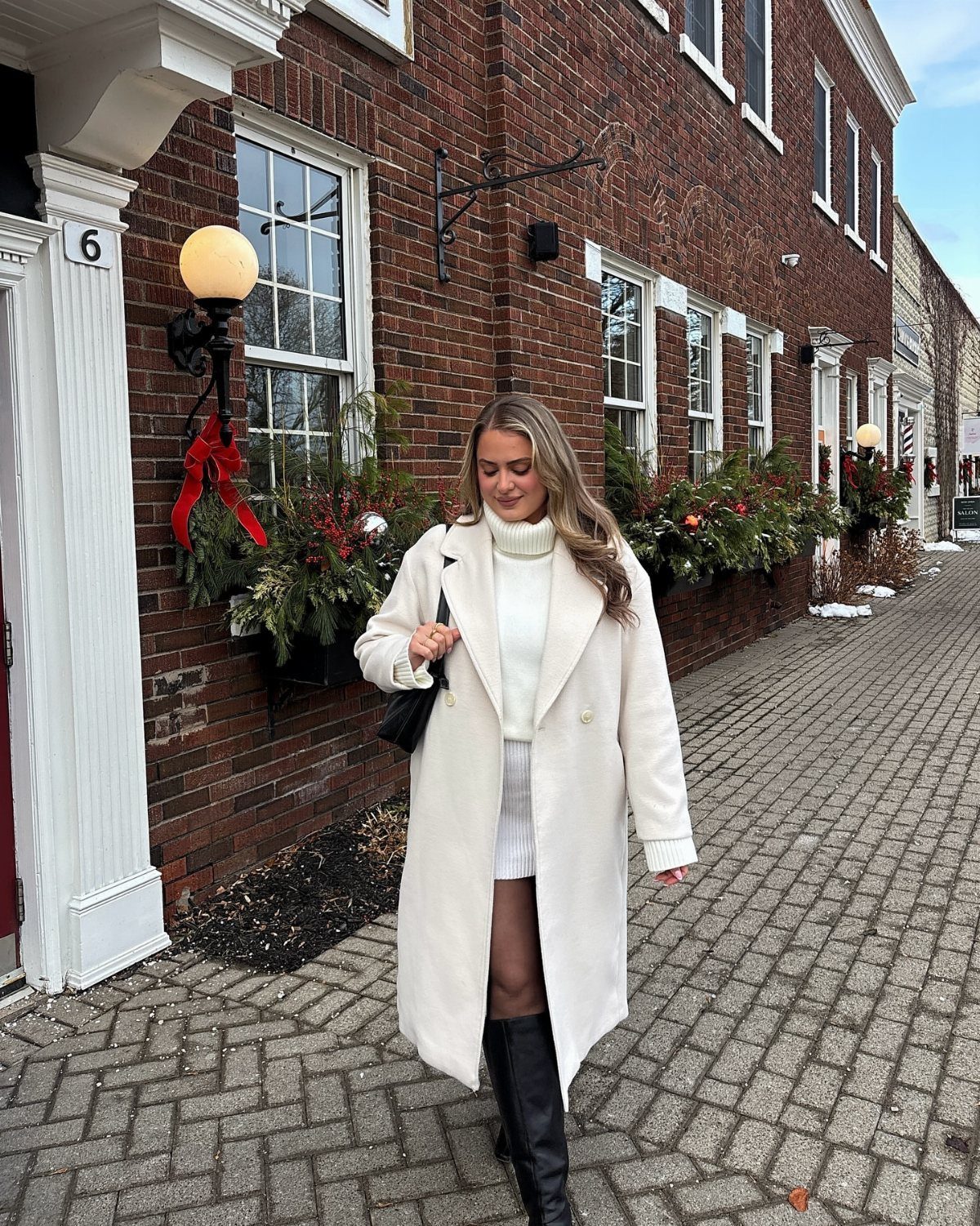 Woman in a white coat, white sweater, and white skirt walking on a brick sidewalk.
