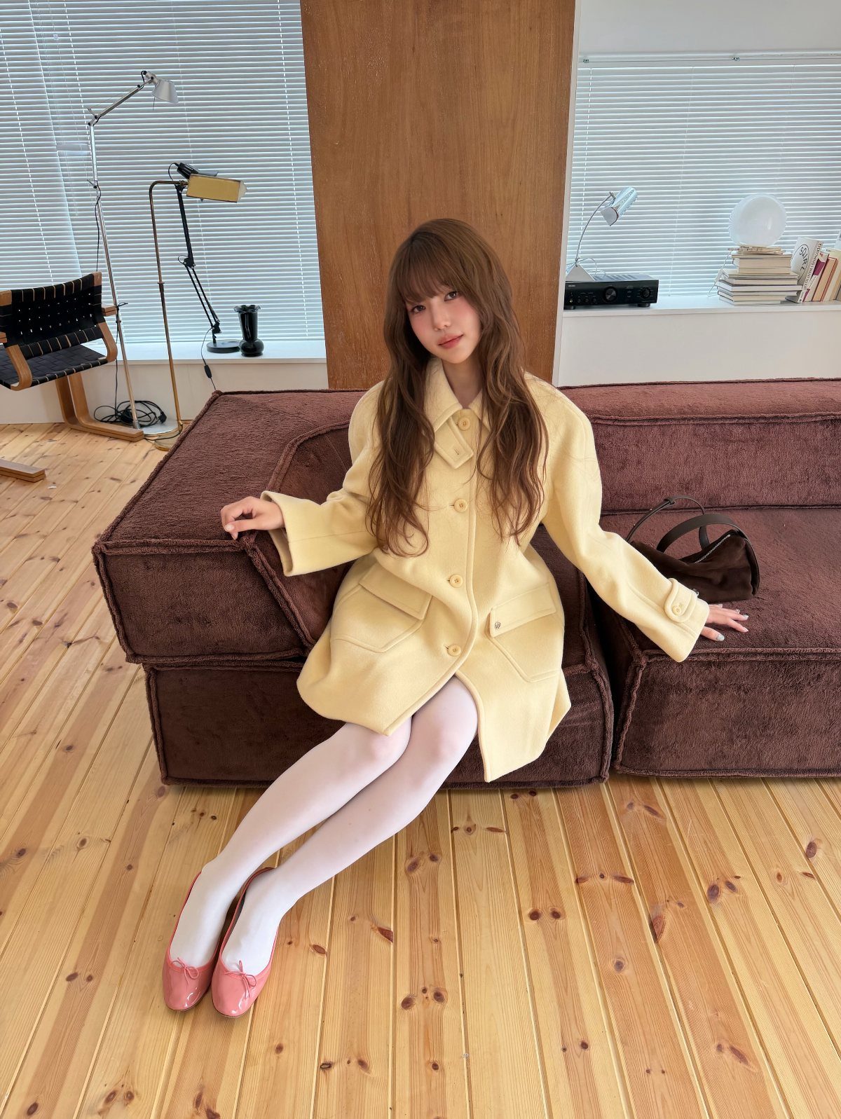 A woman wearing a pastel yellow coat, white tights, and pink ballet flats sitting on a sofa.