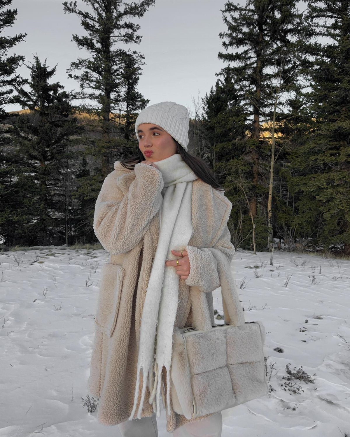Woman in a beige sherpa coat and white knit accessories standing in a snowy forest.