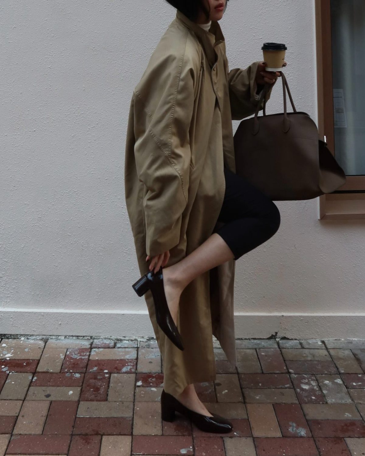 Person in a long tan trench coat, black leggings, and black low-heeled pumps holding coffee.