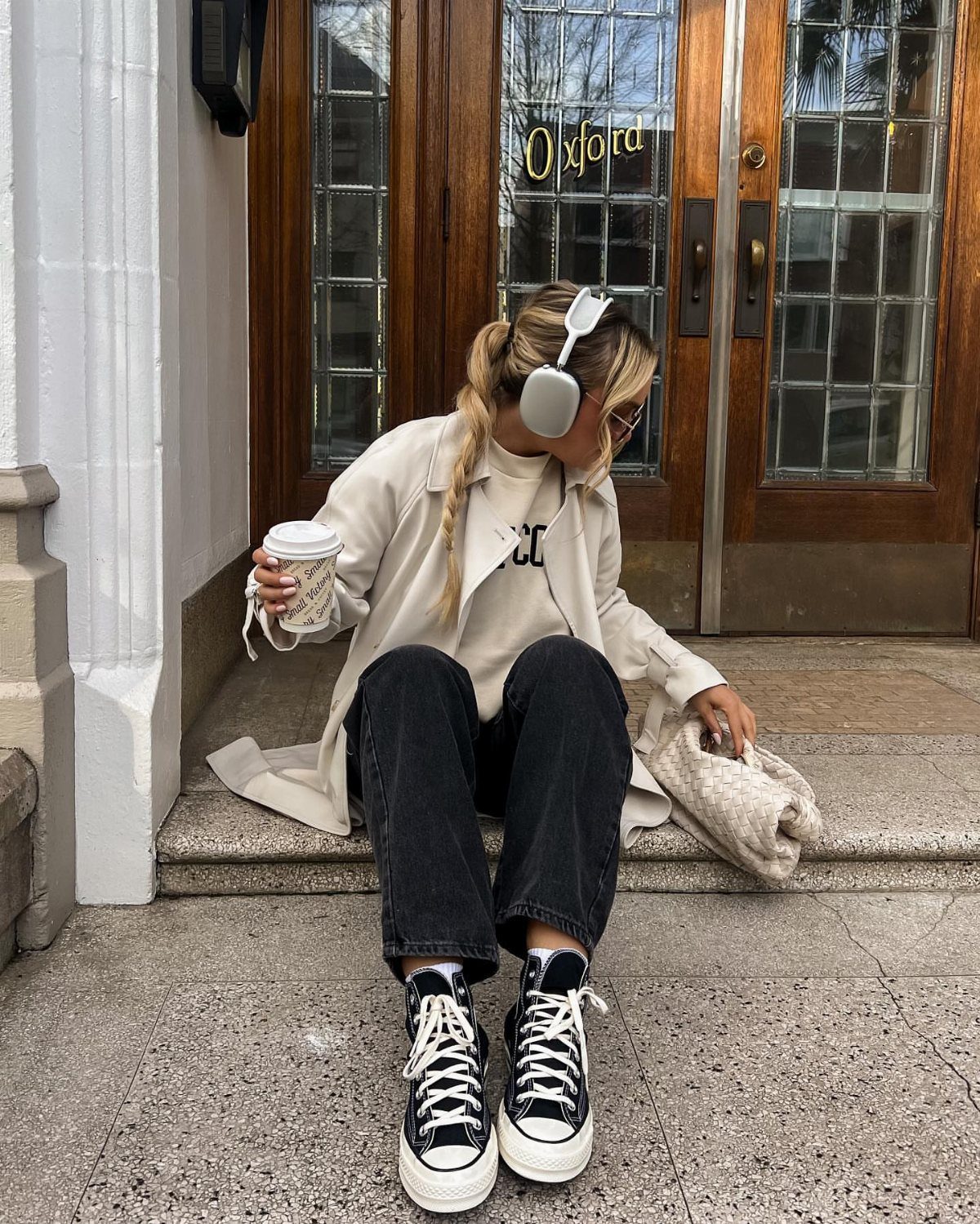 Person sitting on steps wearing black trousers, a cream trench coat, and black Converse sneakers.