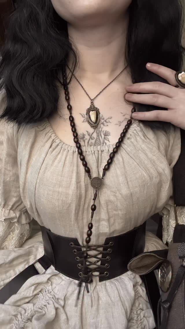 Person wearing a cream linen blouse, black lace-up corset, and dark beaded necklaces.