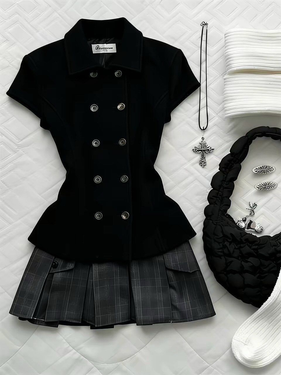 A black blazer and grey plaid skirt laid out with a quilted bag and silver cross necklace.