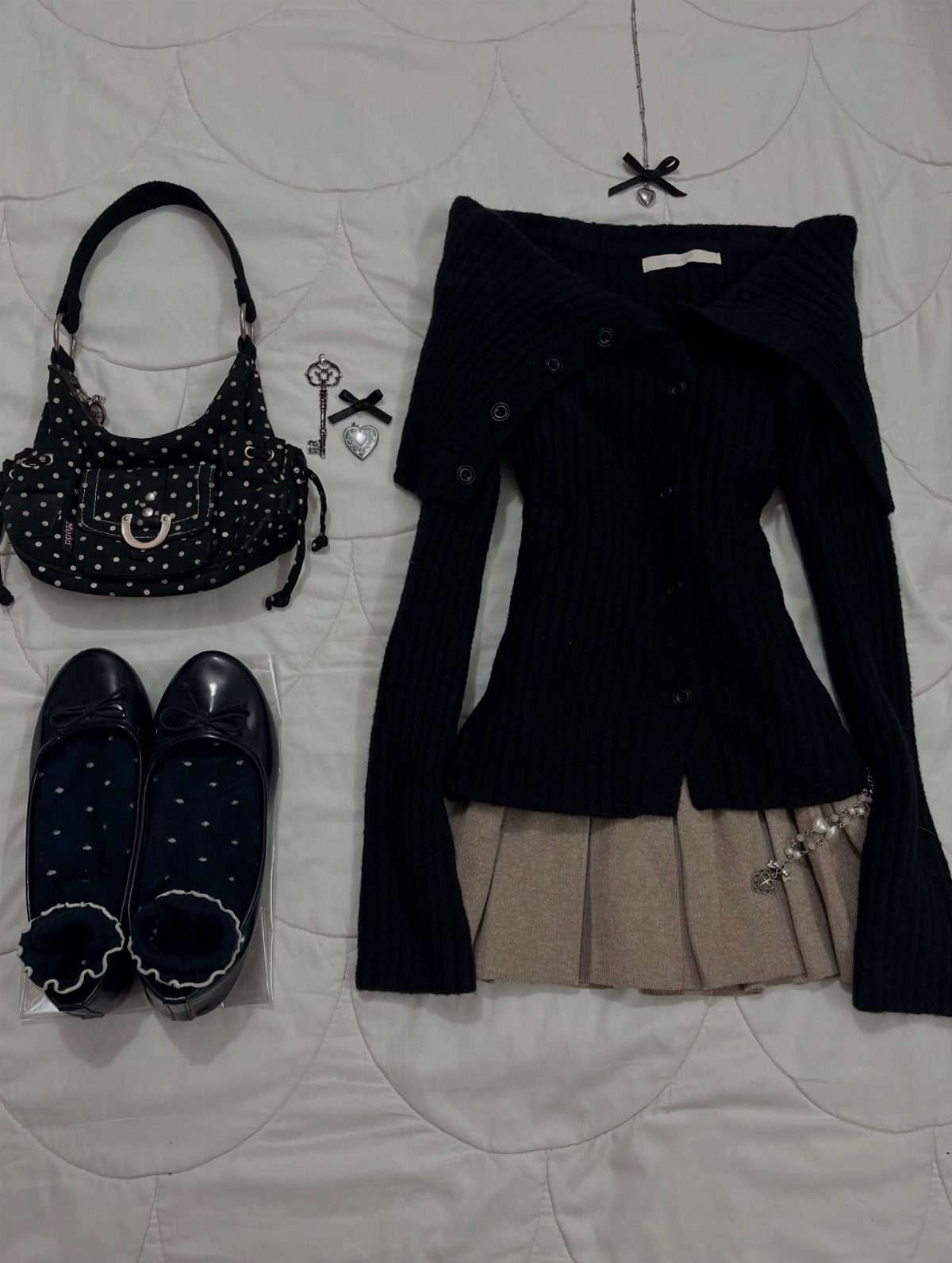 A dark coquette outfit flat lay with a black cardigan, tan skirt, polka dot bag, and loafers.