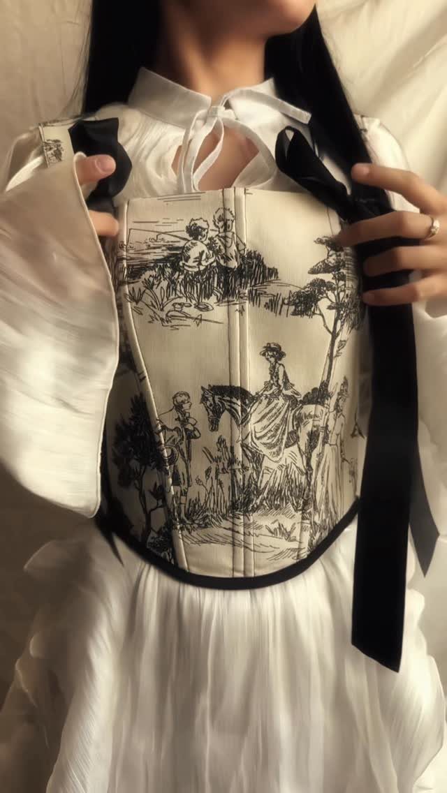 A person wearing a white blouse and a cream corset with black toile de jouy patterns.