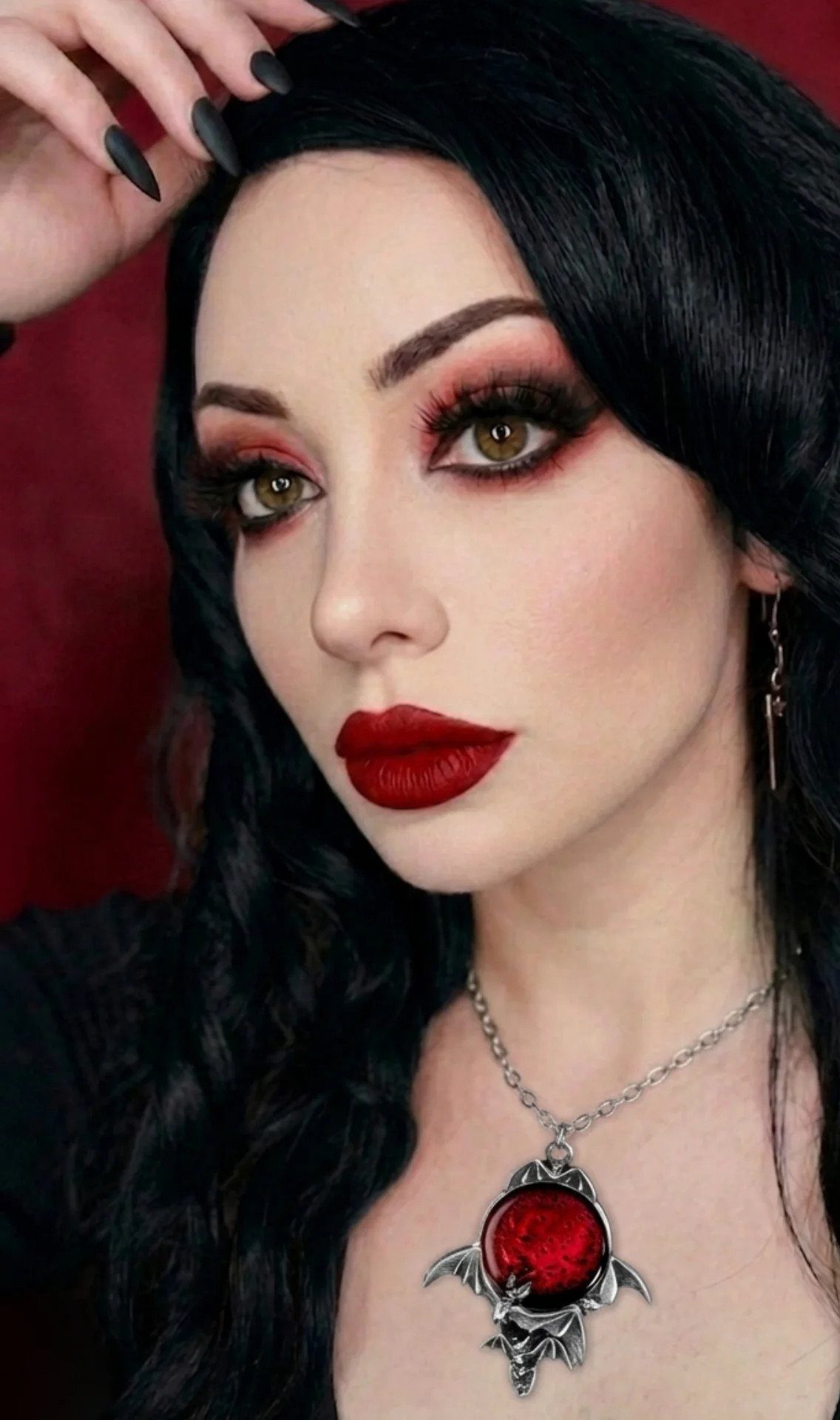 Woman with dark smokey eye makeup, deep red lips, and long black hair wearing a red gemstone necklace.