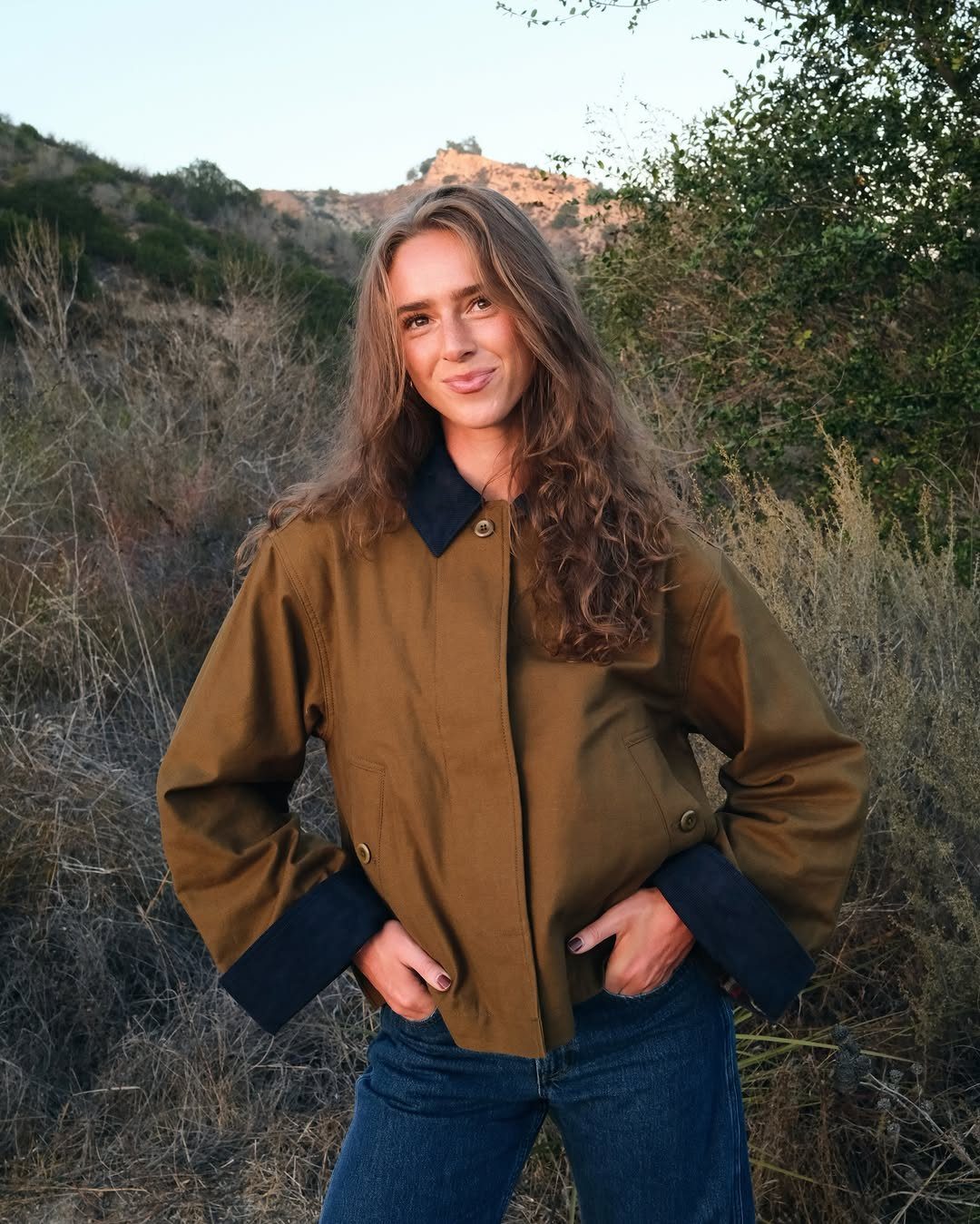 A woman wearing a brown barn jacket and blue jeans standing in a grassy field.