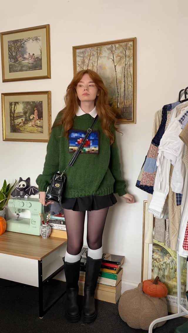Red-haired woman wearing a green knit sweater, black skirt, tights, and black boots.