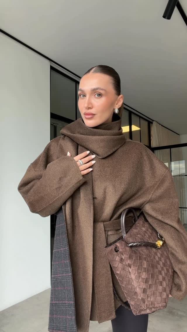 A woman wearing a large, oversized brown scarf coat and holding a patterned handbag.