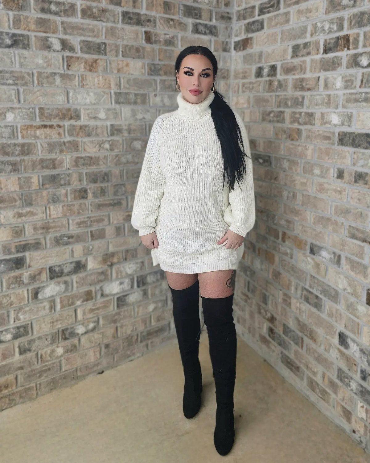 Woman wearing a white knit sweaterdress and black over-the-knee boots against a brick wall.