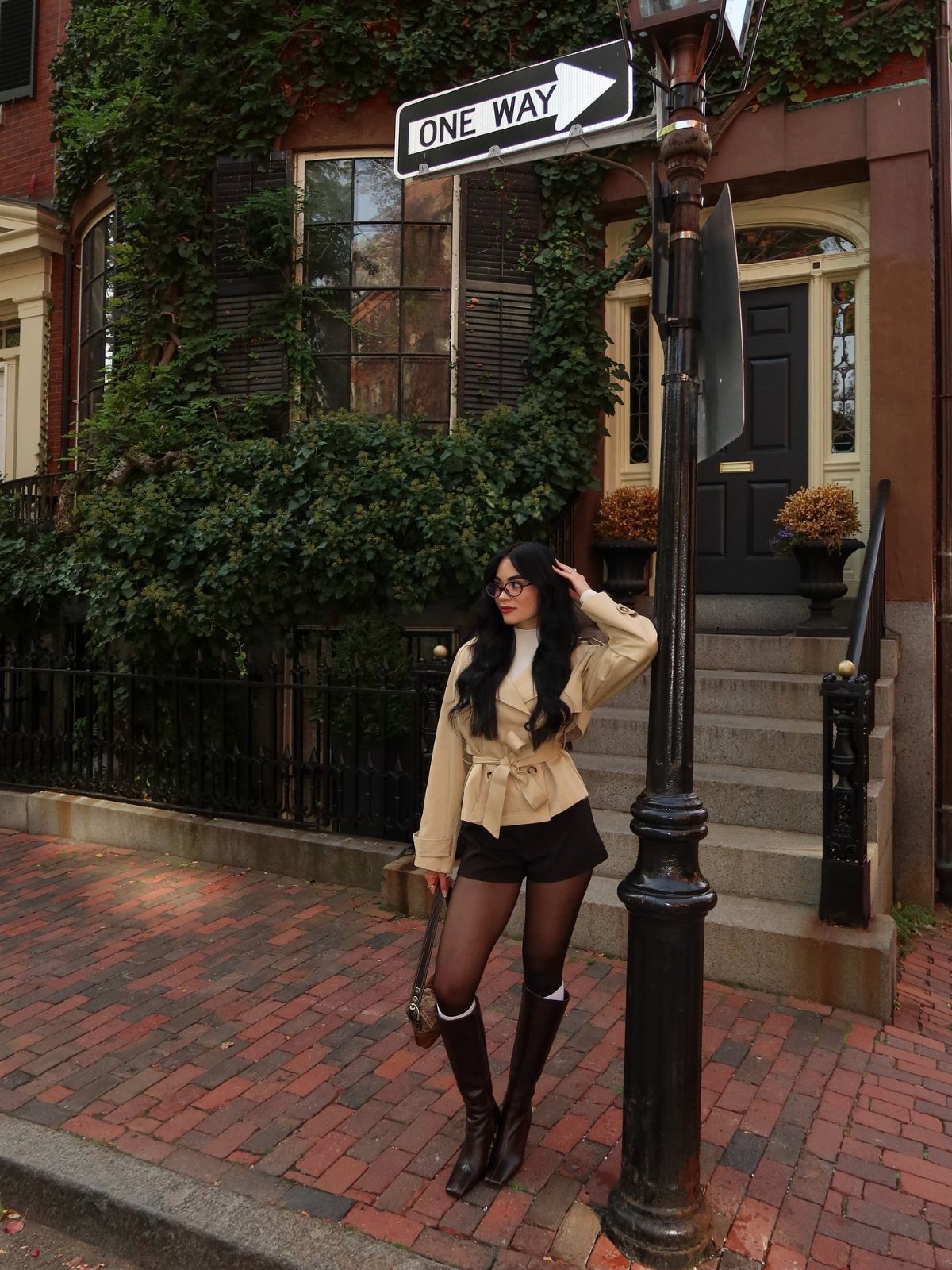 A woman in a tan trench coat, white turtleneck, and tall boots posing on a brick street.
