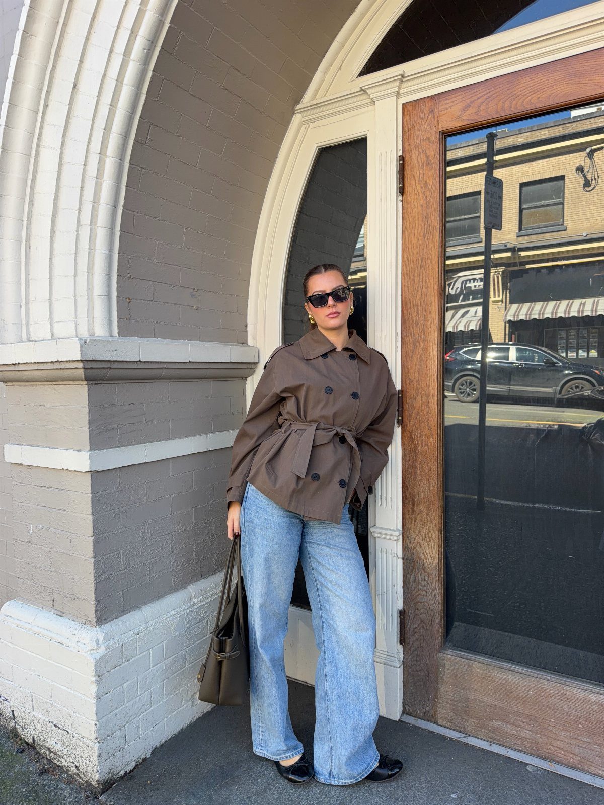 Woman in a brown trench coat and wide-leg blue jeans standing by an arched doorway.
