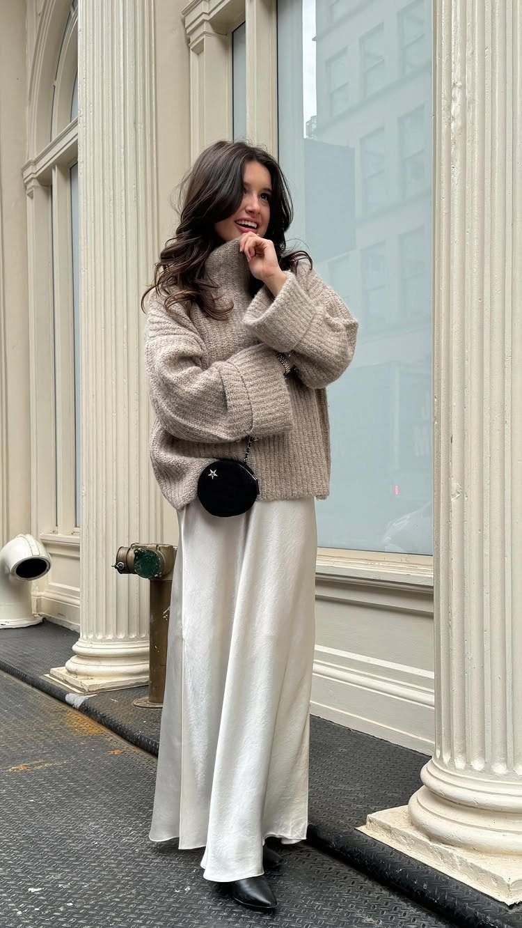 Woman wearing a beige turtleneck sweater and long white satin skirt standing by columns.