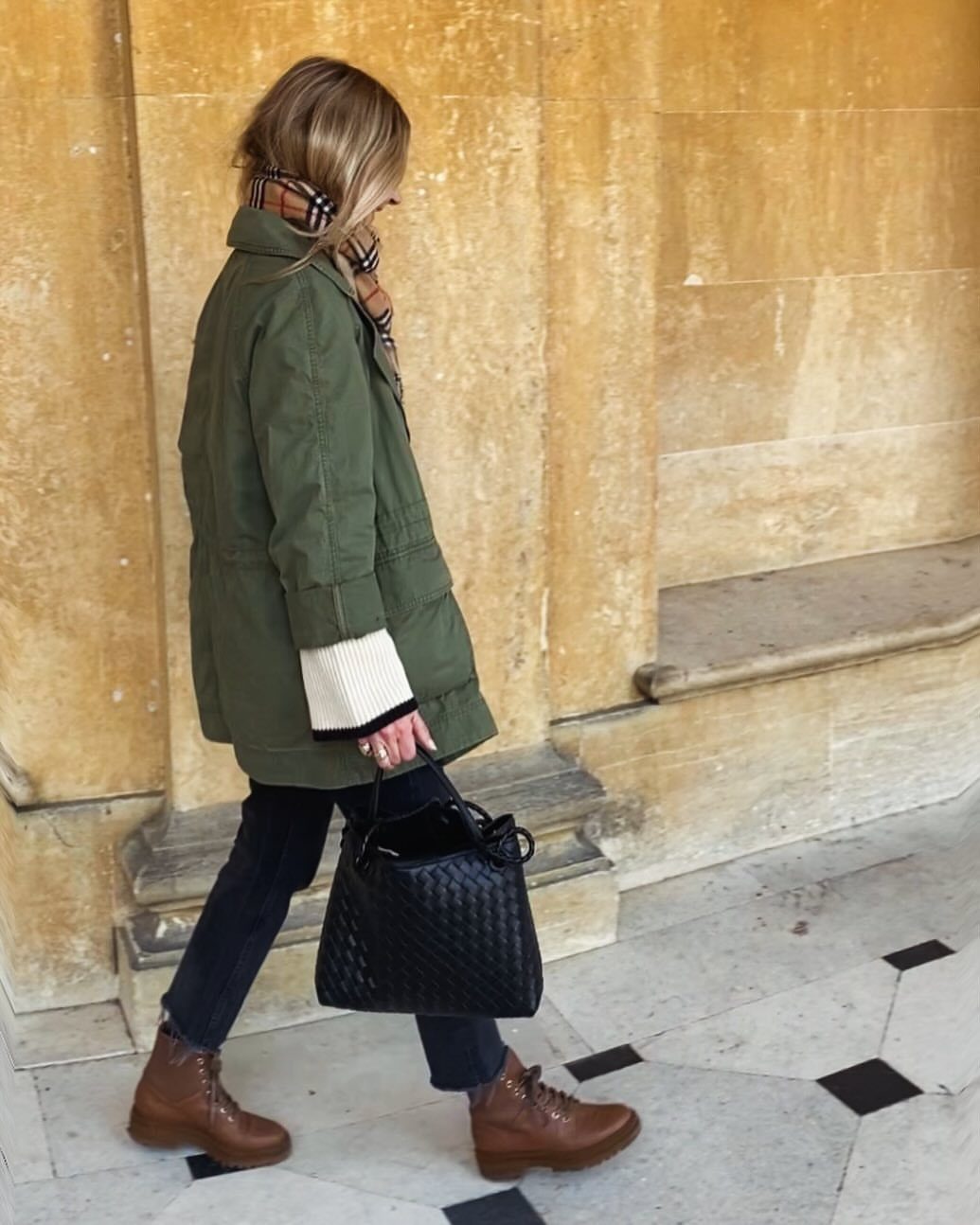 A woman walking in an olive green utility jacket, dark jeans, and brown boots while carrying a black bag.