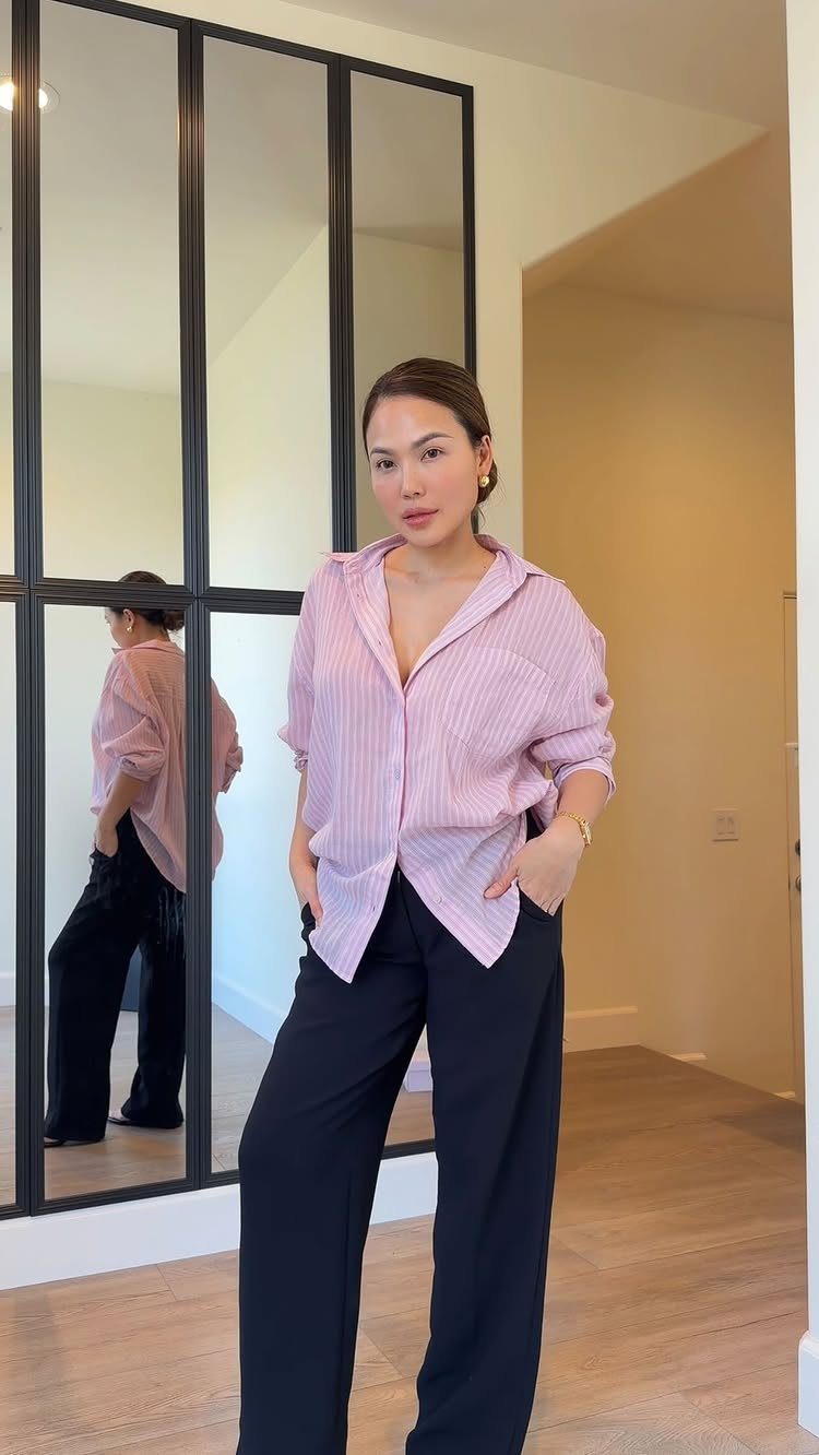 A woman wearing a pink striped shirt and black wide-leg trousers posing indoors.