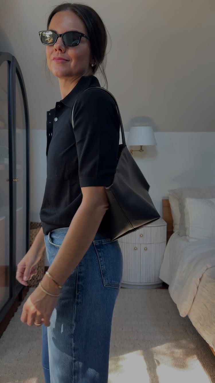 Woman in a black polo shirt and blue jeans wearing sunglasses and carrying a handbag.