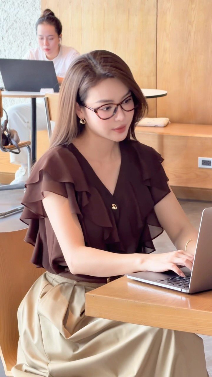Woman in a ruffled brown blouse and satin skirt working on a laptop at a wooden desk.