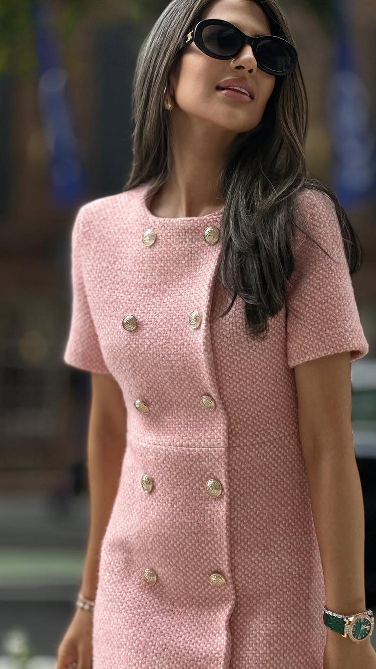 A woman wearing a pink tweed dress with gold buttons and black sunglasses.