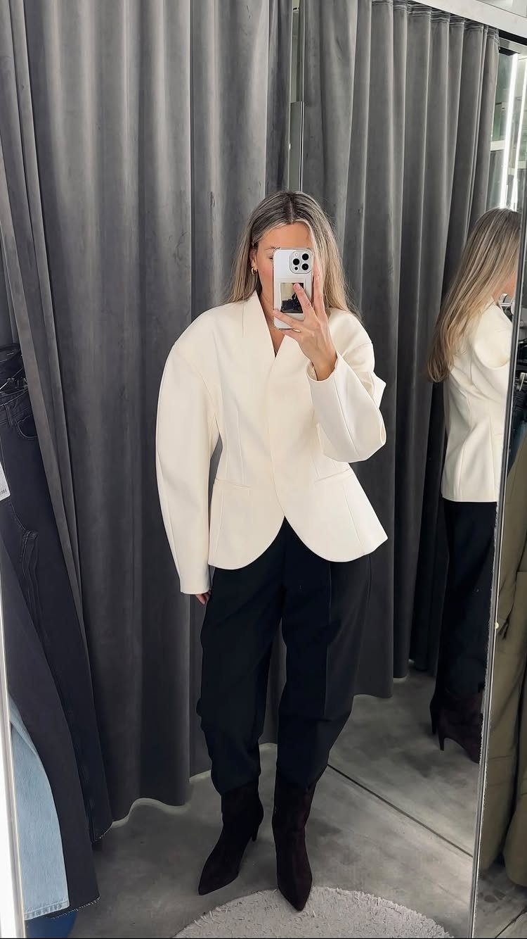 A woman posing in a mirror wearing a white tailored jacket and black trousers.