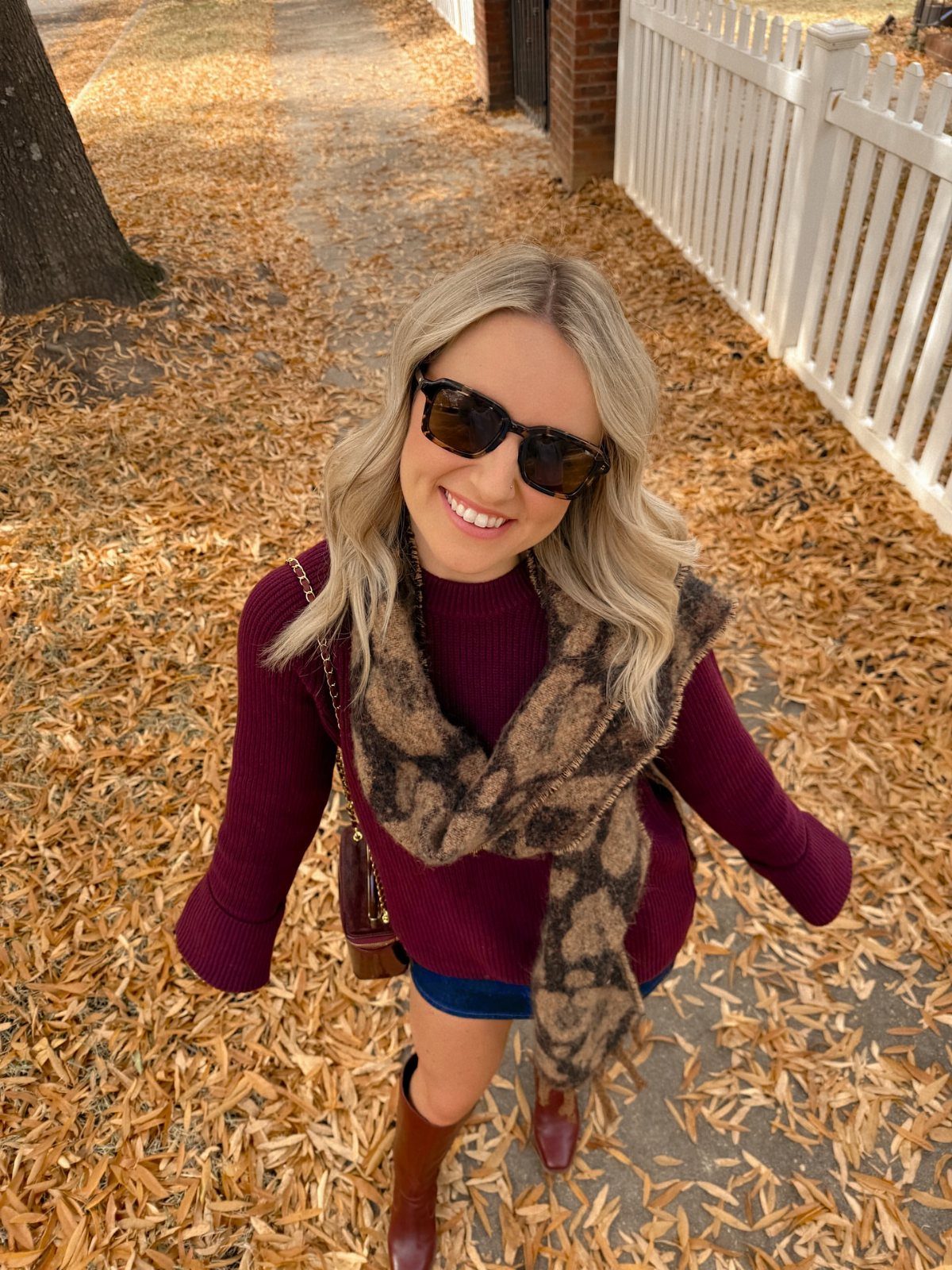 Smiling blonde woman in a burgundy sweater and tall brown boots standing on autumn leaves.