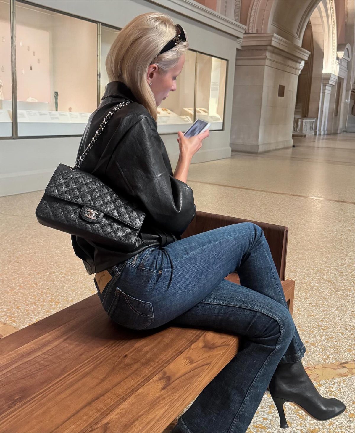 A woman in a leather jacket, blue jeans, and black ankle boots sitting on a bench.