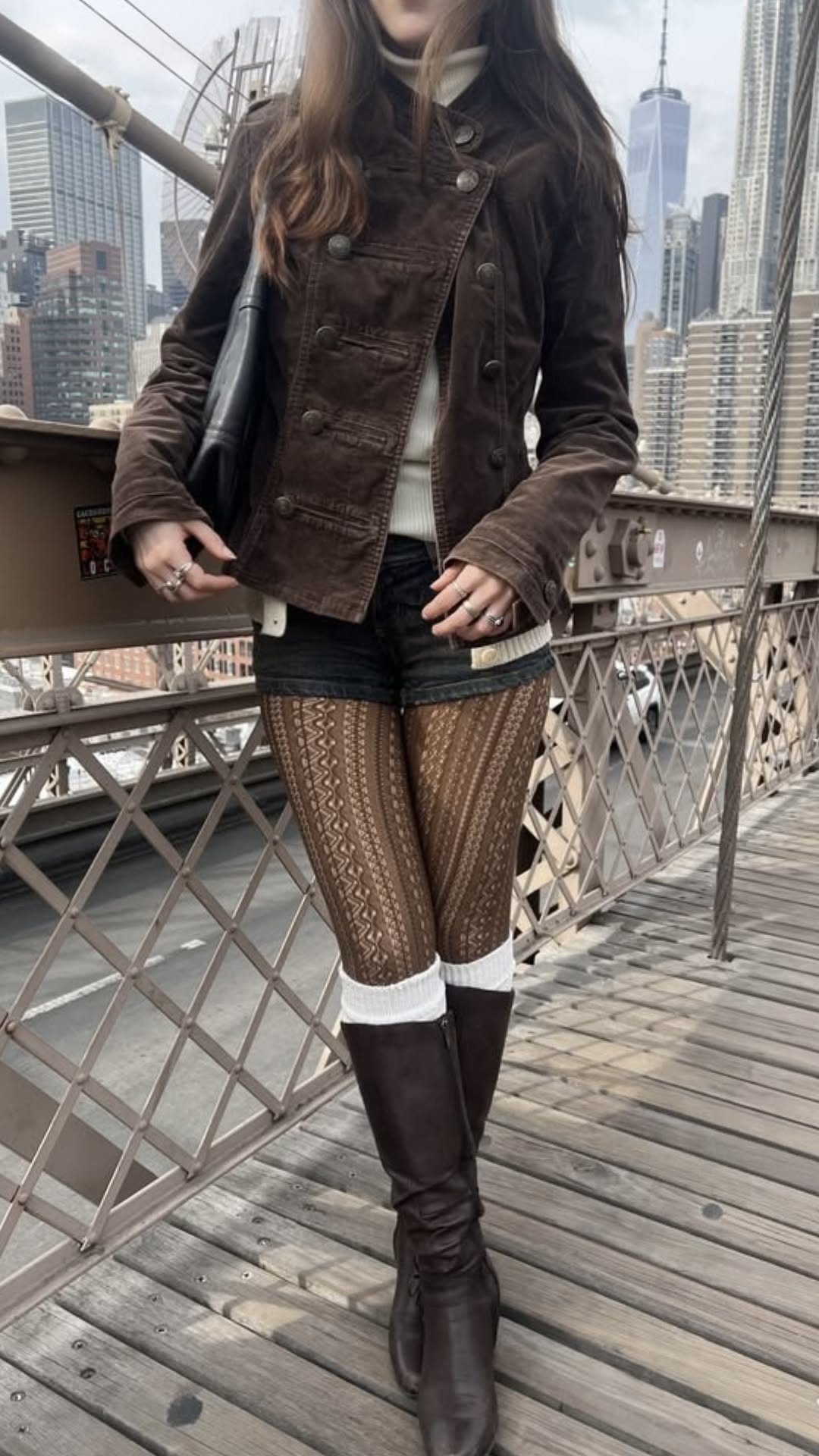 A person wearing a brown jacket, patterned tights, and tall brown boots on a city bridge.