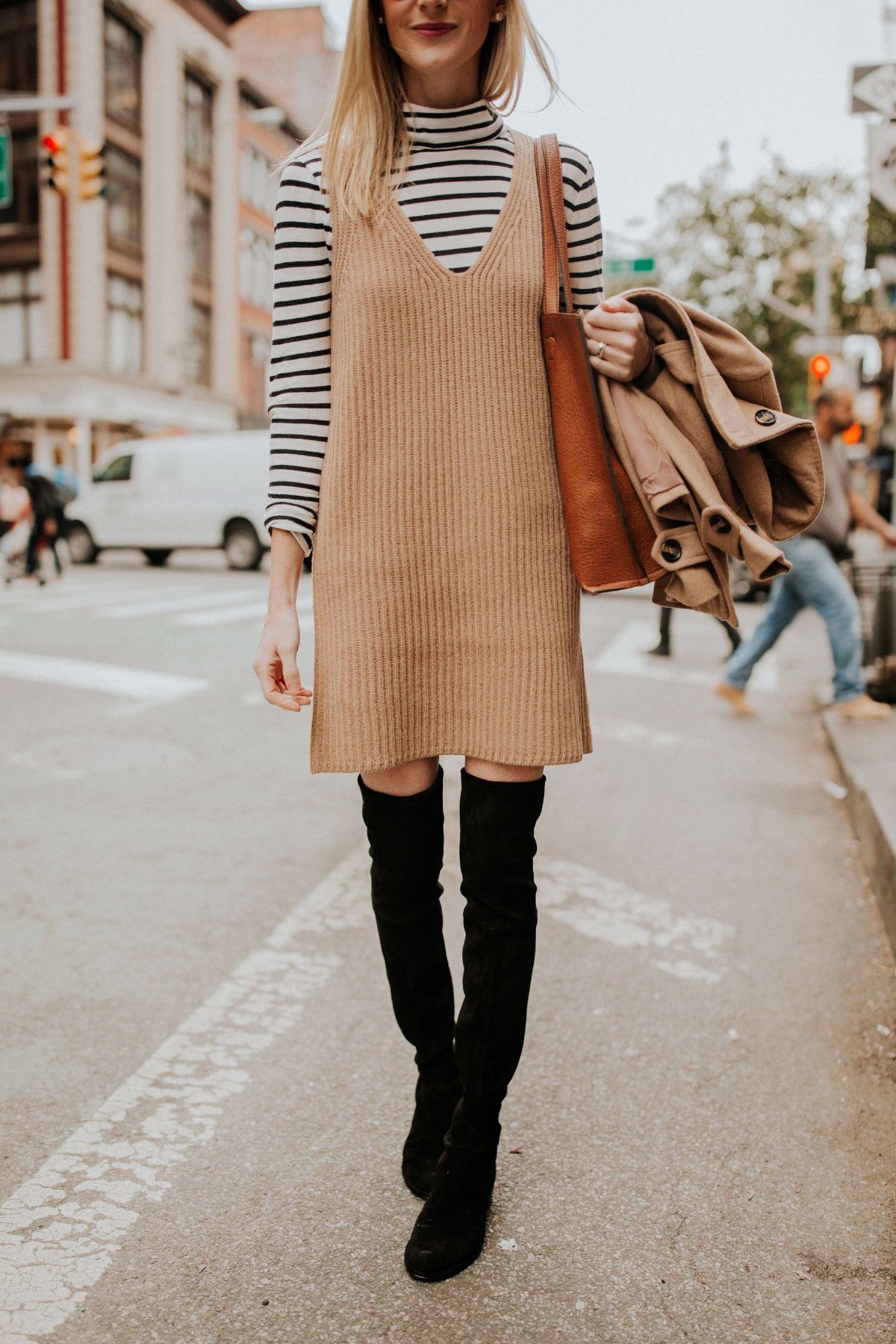 Woman wearing a tan sweater dress and black over-the-knee boots walking on a city street.