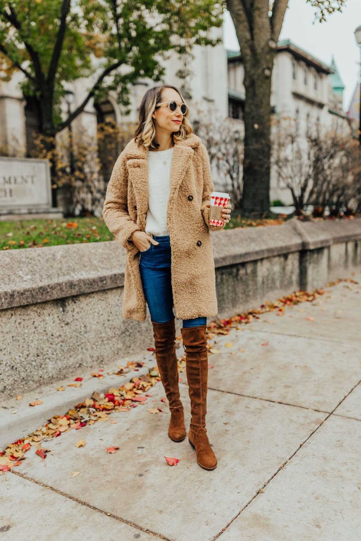Woman wearing a tan teddy coat and brown over-the-knee boots walking outdoors.