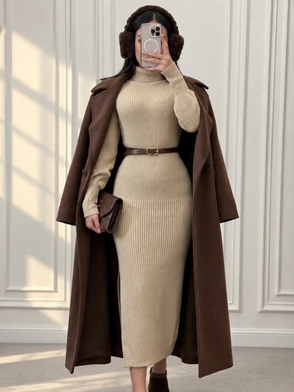 Woman in a beige ribbed knit midi dress and long brown coat posing for a selfie.