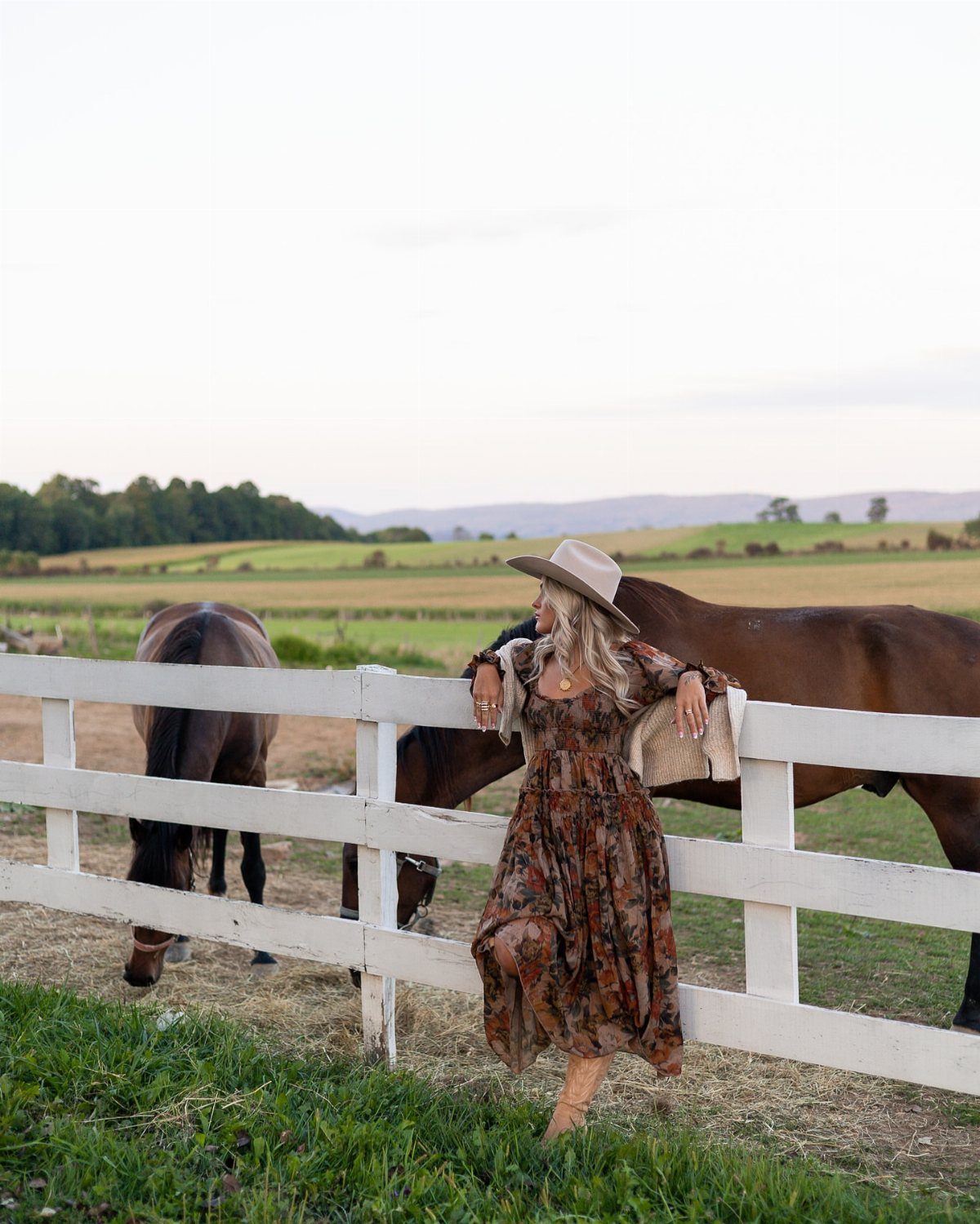A woman in a floral midi dress leans against a white fence in a grassy field.