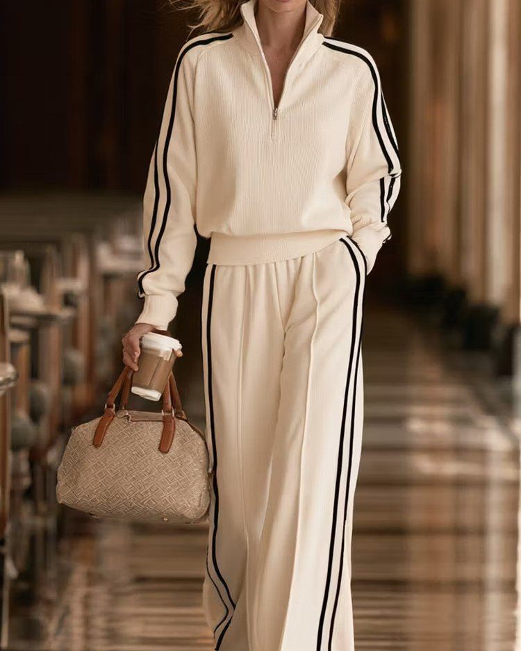 Person wearing a cream tracksuit with black stripes walking while holding a handbag and coffee.