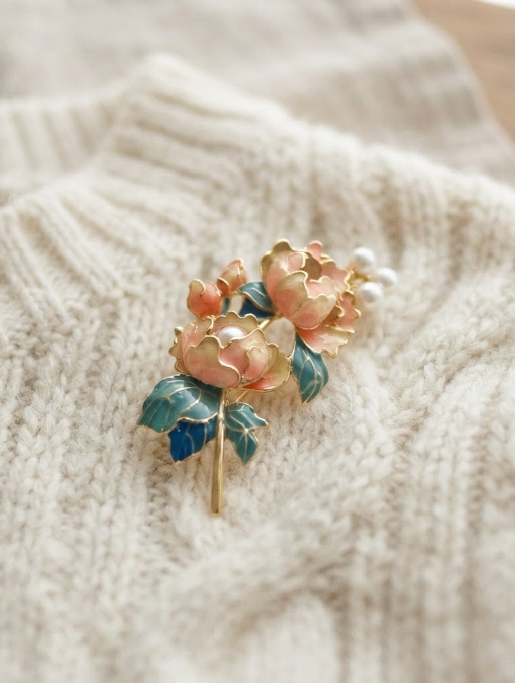 A peach and teal floral brooch pinned to a cream textured knit sweater.