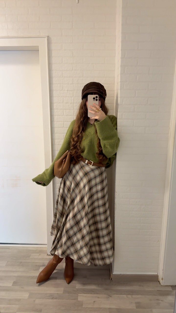 A person in a green sweater and plaid midi skirt posing for a mirror selfie.
