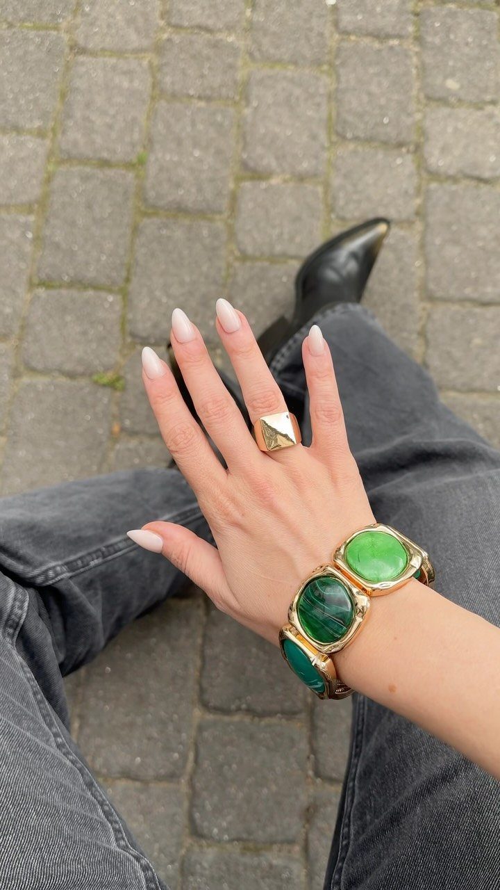 A hand wearing a chunky gold ring and a large green stone bracelet over dark denim jeans.