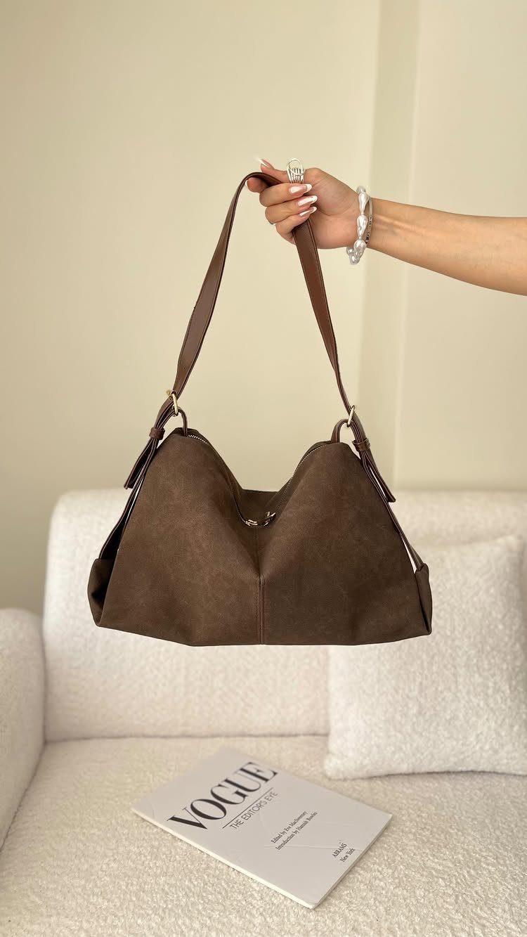 A hand holding a large dark brown suede hobo handbag against a neutral background.
